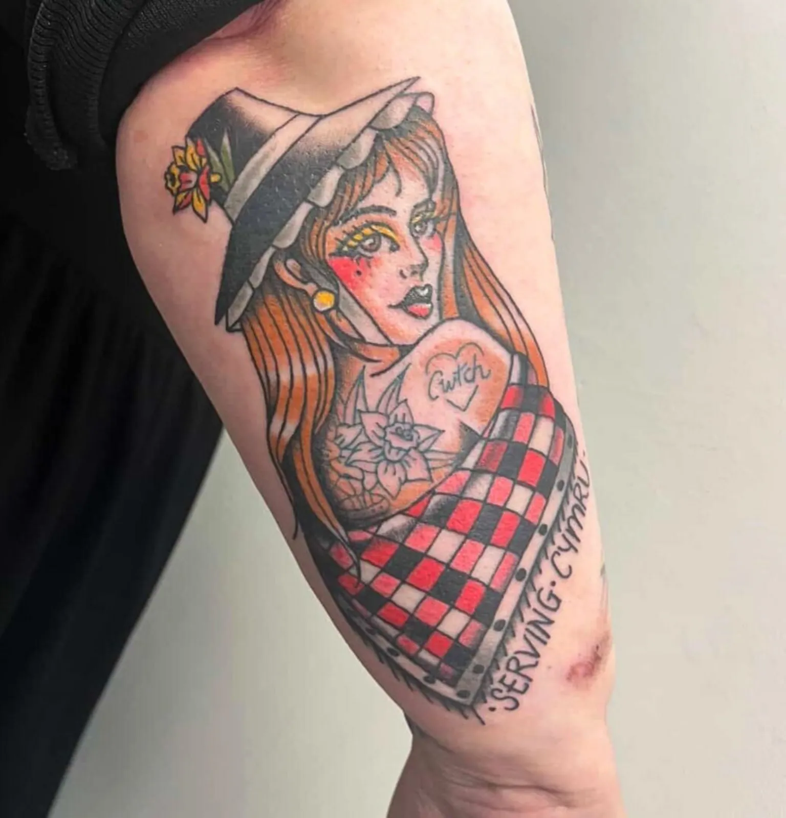 Jessica Tucker A traditional style tattoo on the back of a woman's forearm. The tattoo is of a woman wearing a traditional Welsh chimney hat, with a daffodil on it, and a black white and red shawl. The woman has long red hair and rosy cheeks. She looks back over her shoulder and the pin up has her own tattoos on her back and shoulder of daffodils and a heart with the word