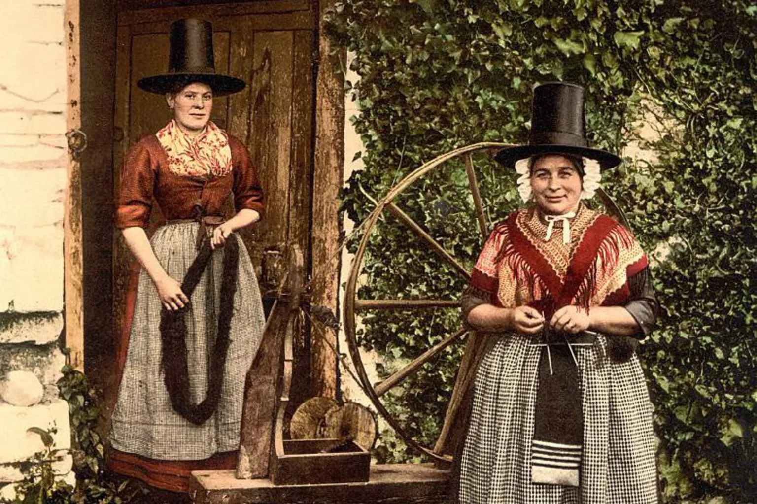 Universal Images Group via Two women stand next to a large wooden spinning wheel. They wear traditional Welsh clothing - a black chimney hat, red shawls, and black and white aprons.
