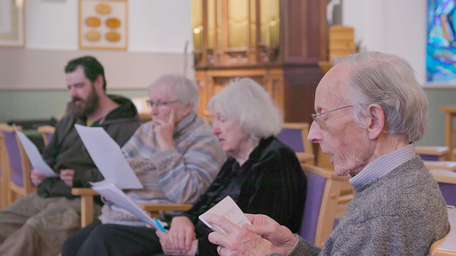 Hopscotch A group of people singing hymns. There are four of them in total and they are seated and reading from a sheet of paper. The man closest to the camera has grey hair and glasses and a grey jumper. Next to him is a grey-haired woman in a black top, then a grey-haired woman with glasses and a striped jumper and finally a man with black hair and a beard. They are in a church setting.