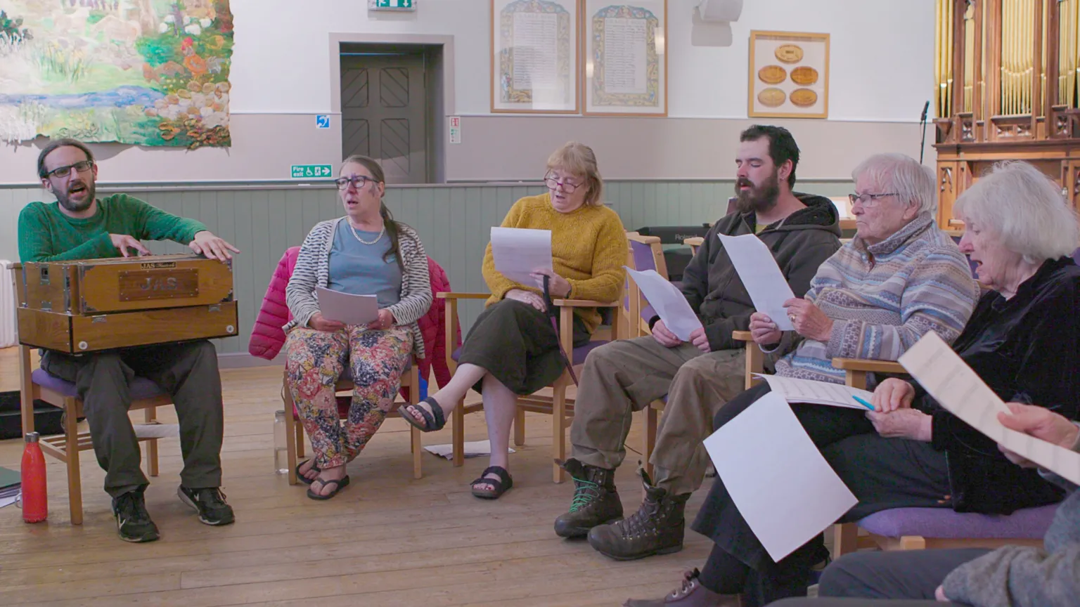 Hopscotch A man in a beard and glasses leads five other people - four women and a man - in song. They look like there is an organ behind them and they are all holding sheets of paper apart from the man leading them who appears to be playing some kind of musical instrument, a wooden box on his knees.