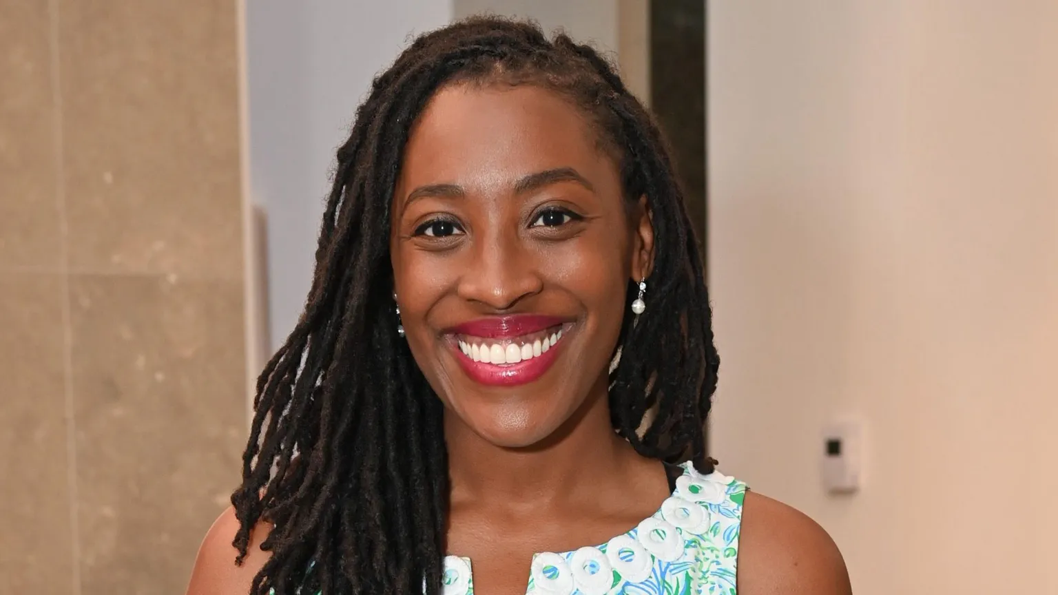 Dr Imani Strong Dr Imani Strong is smiling at the camera. She has shoulder-length dark hair and is stood up against a granite tile wall and a white wall in the distance. She is wearing a white, blue and green patterned dress.