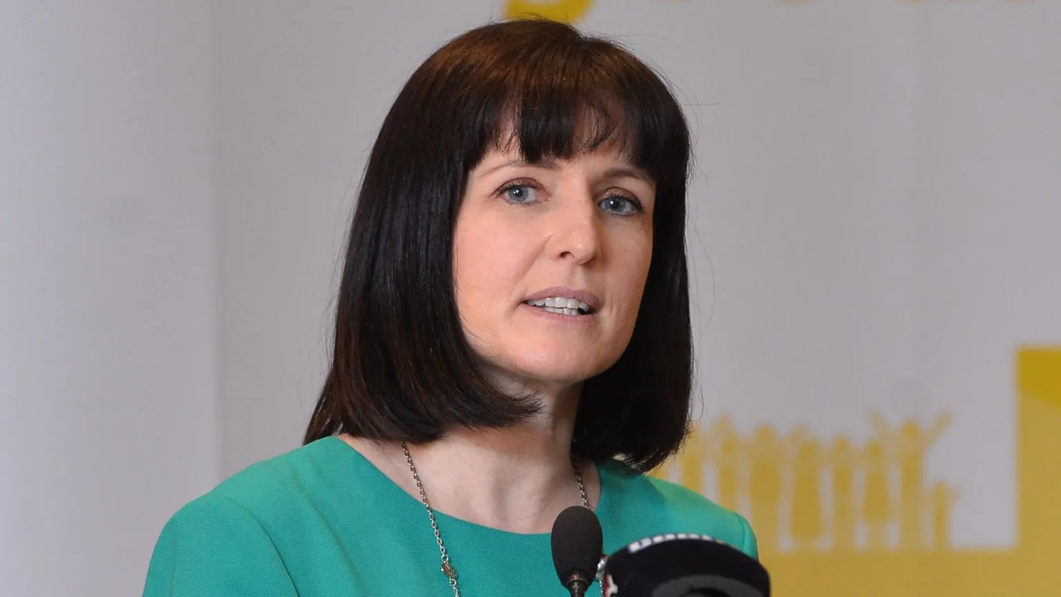 Pacemaker Alliance assembly member Paula Bradshaw, she had dark shoulder length hair and has a fringe. She is wearing a green dress and is speaking into a microphone.