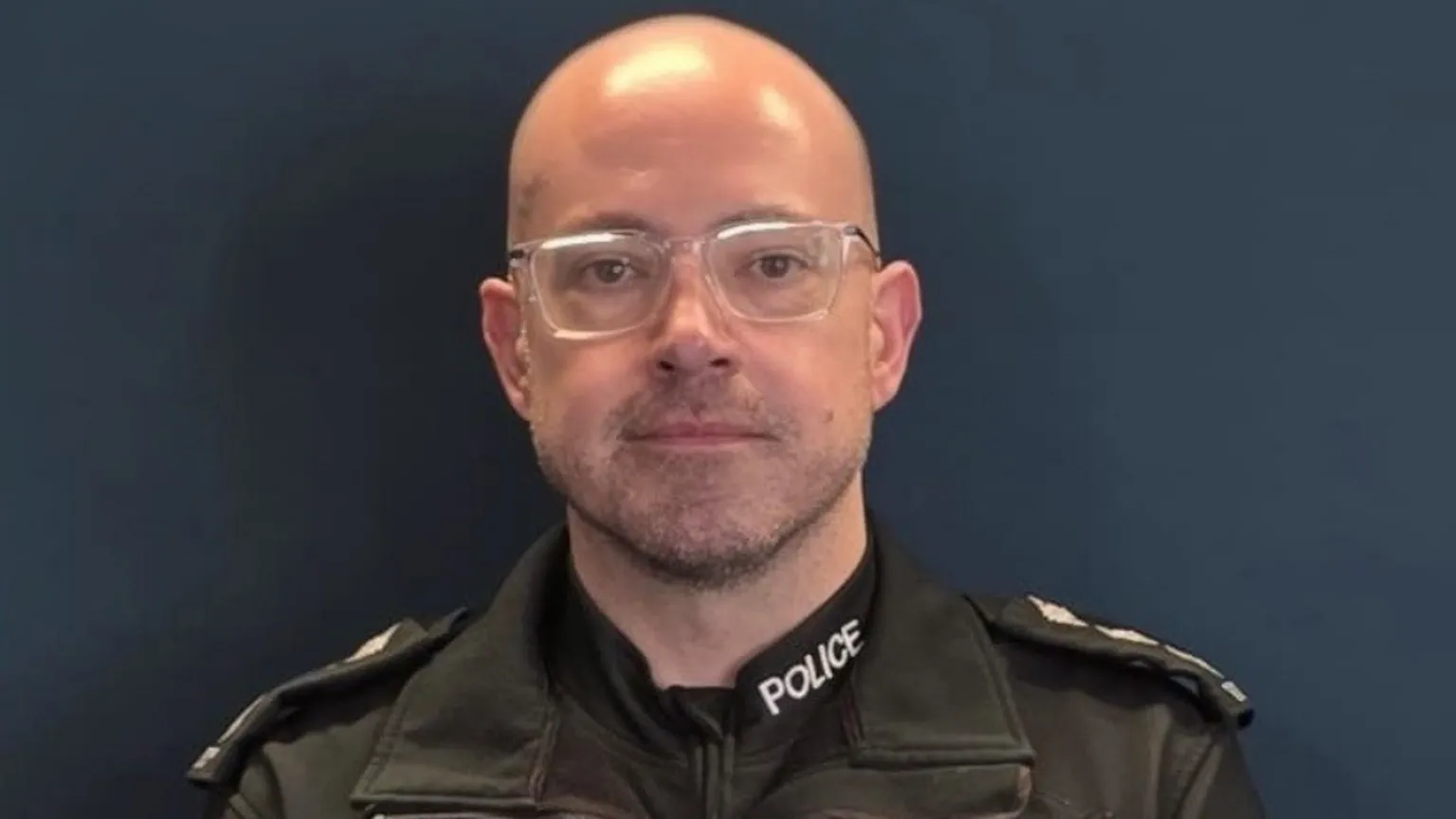 Police Service of Northern Ireland Paddy Mullan is a middle-aged bald man. He is looking at the camera with a neutral expression on his face. He is wearing a pair of clear framed glasses and a police uniform. He is stood against a plain, dark wall.