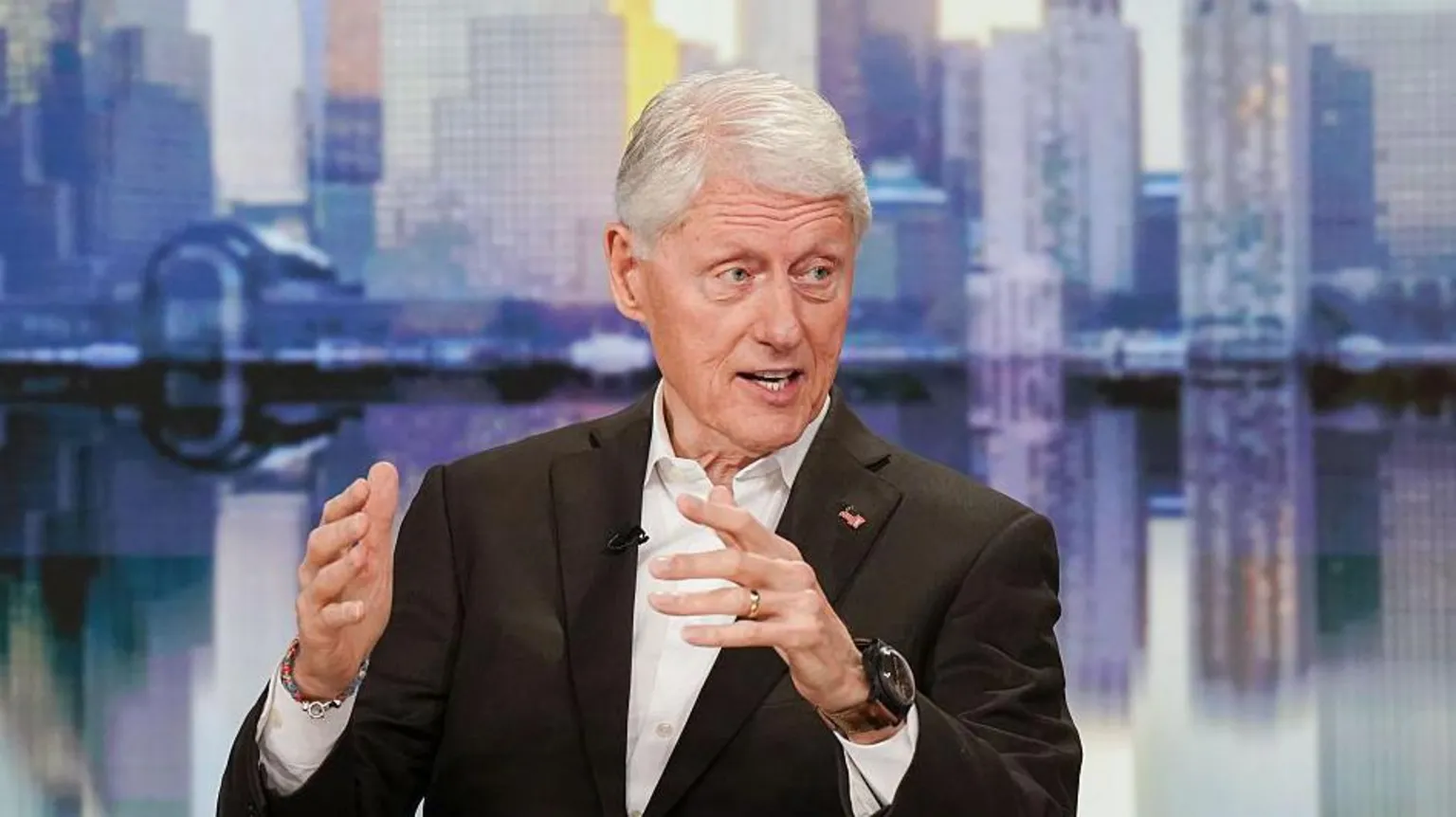  Bill Clinton wears a black suit and a white shirt on a talk show tv set that has backdrop of New York City