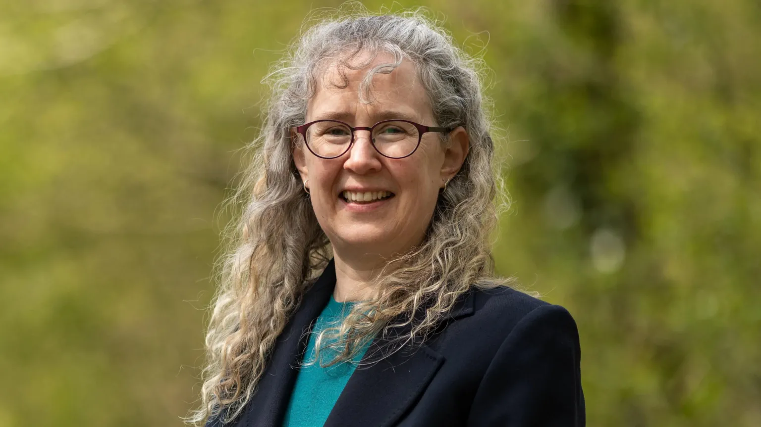 Royal Society for the Protection of Birds NI (RSBP NI) Dr Sharon Thompson is pictured standing in front of some trees but they are blurred. She is a middle aged woman with grey, curly hair. She is smiling at the camera and is wearing a pair of circular glasses, a navy blazer and a teal shirt.