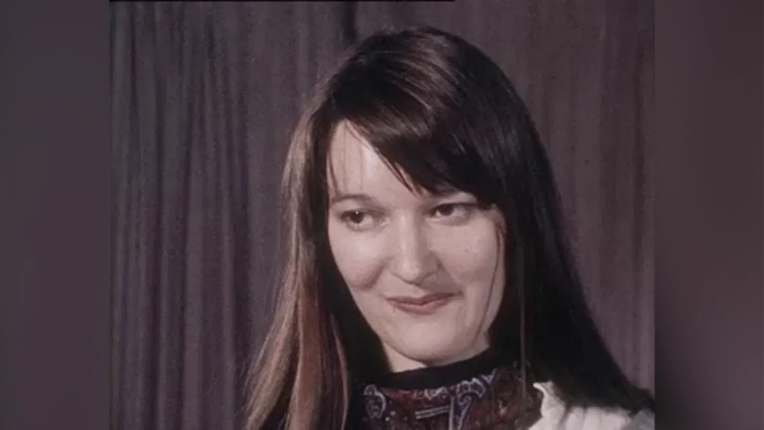 Elizabeth McCrum is smiling. Her long brown hair is down and she has a side fringe. She is wearing a scarf. 