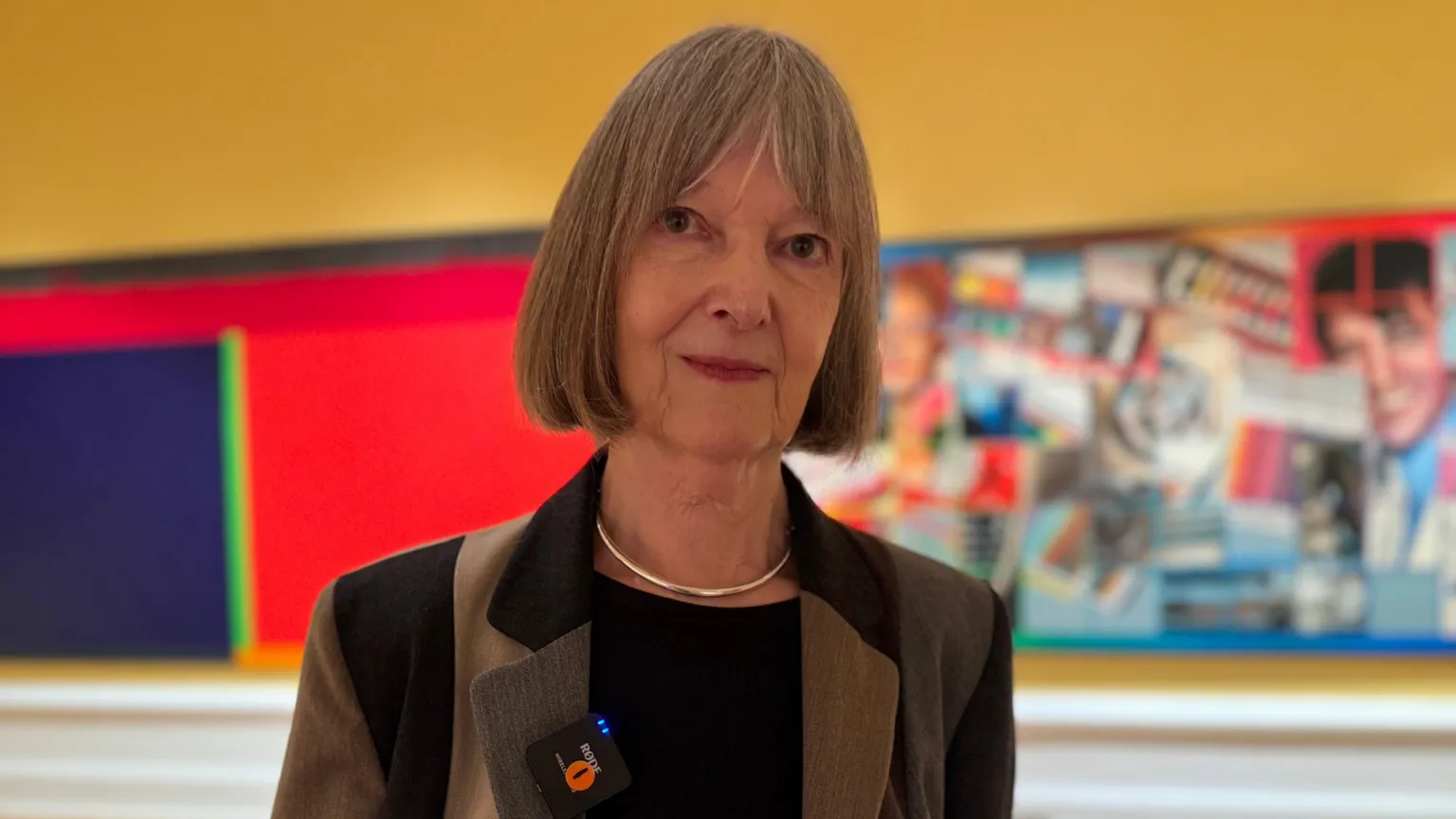 Elizabeth McCrum is wearing a brown and beige jacket. She has bobbed hair. She has a silver necklace on. Paintings are behind her. 