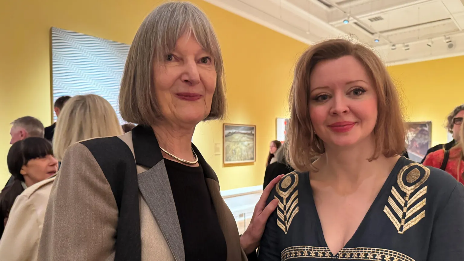 Elizabeth McCrum and Charlotte McReynolds are standing next to each other. Elizabeth has a hand resting on Charlotte's shoulder. People and paintings are in the background. 