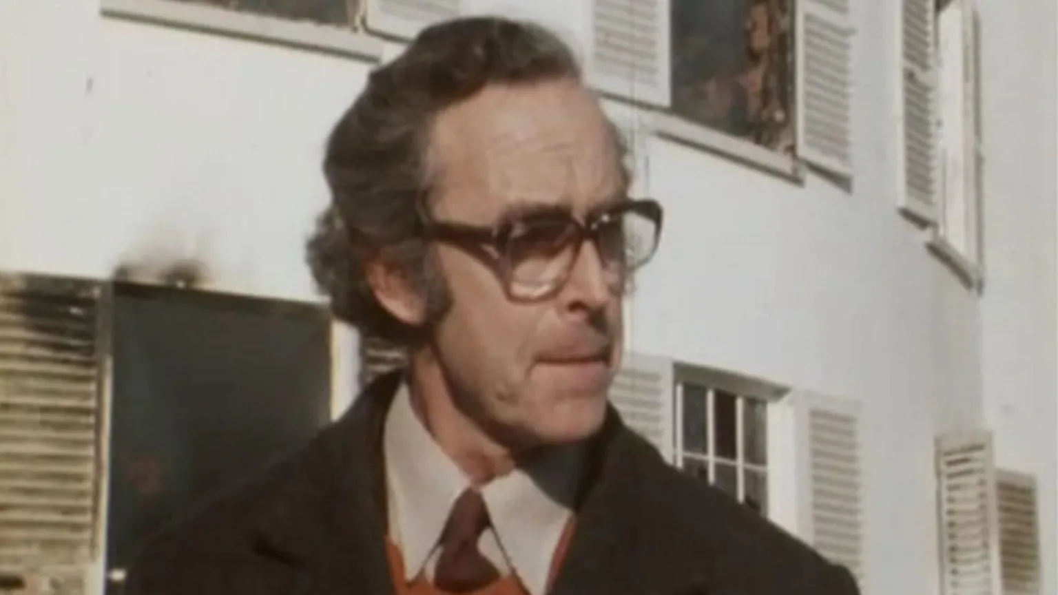 A man is standing outside a building that's on fire. A fire can be seen from one of the windows. He is wearing glasses and a coat, a brown tie with a beige shirt and an orange jumper