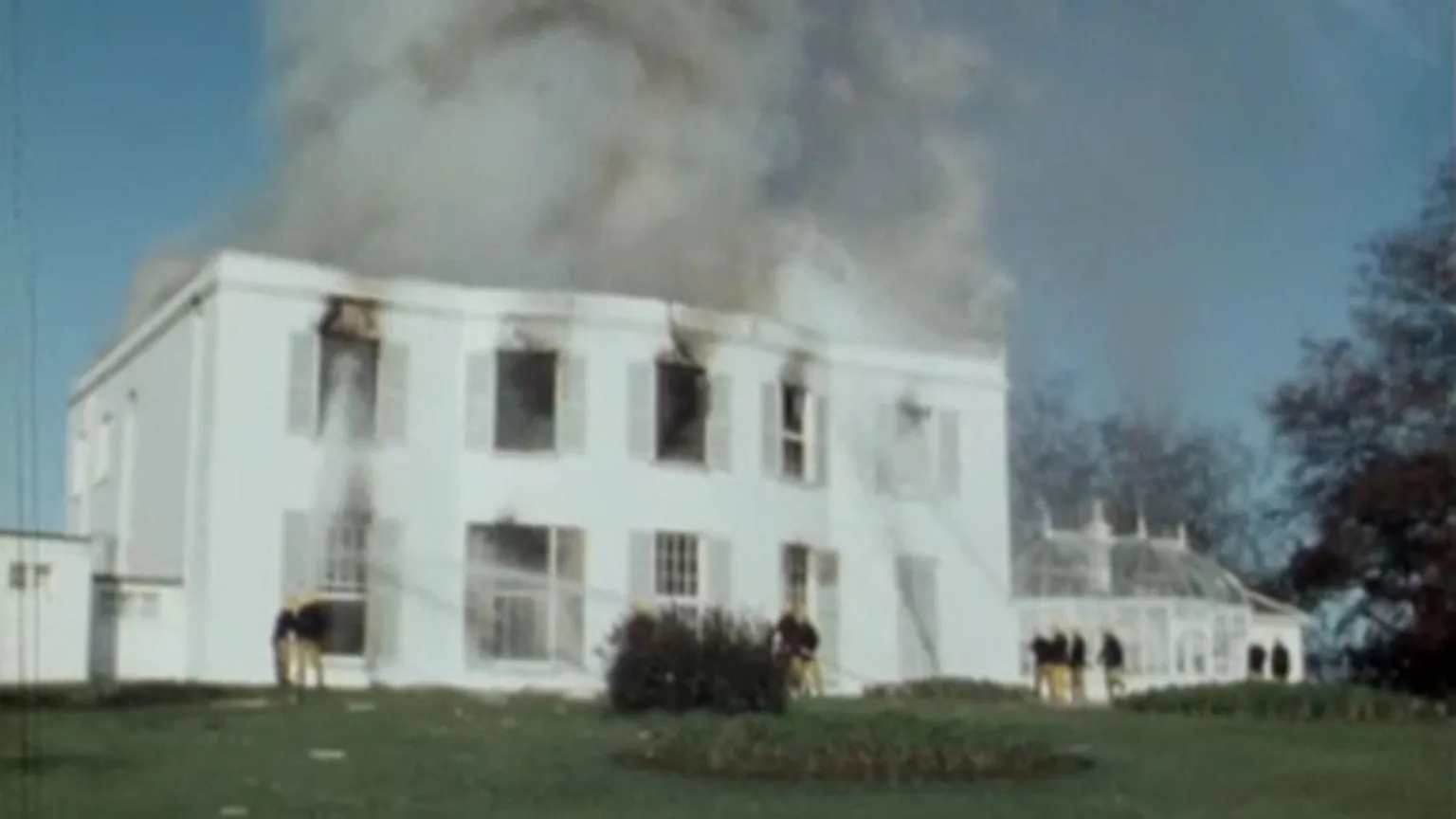 Smoke is bellowing out of a building that's on fire. The 19th century building is white with big windows and a hothouse on the side. Firefighters are close to the building. 