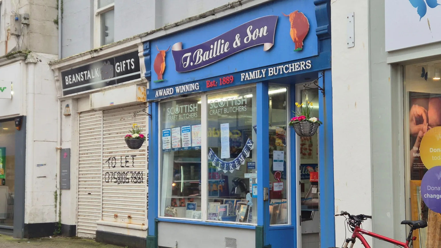 The T Baillie and Son butcher shop - it is a small shop with blue storefront. Bunting saying Retirement is hanging in the window.