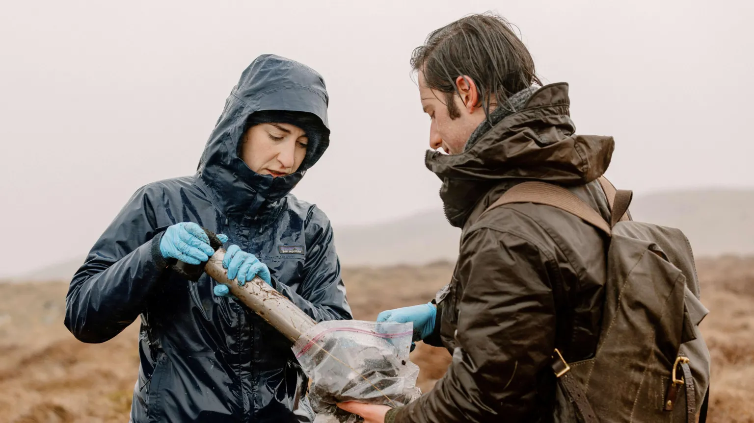 Spun/Seth Carnill Woman wearing a dark raincoat and blue gloves collecting a sample of soil with a man with brown hair wearing a brown coat and rucksack both stood on open grassland.