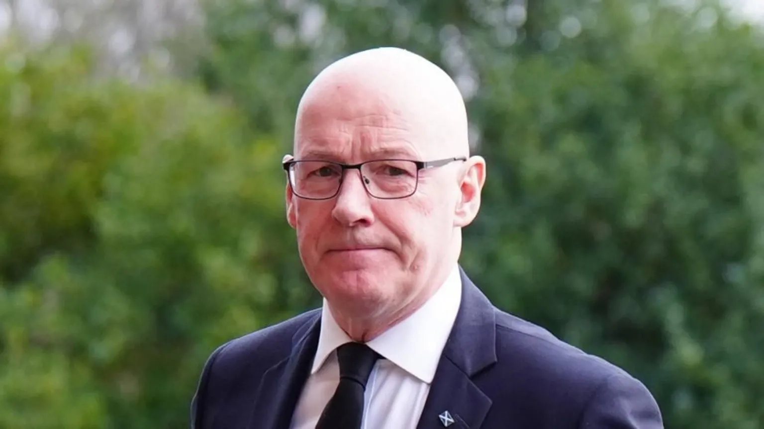 John Swinney, who has a bald head and glasses, is looking at the camera. H is wearing a dark suit with a white shirt and dark tie.