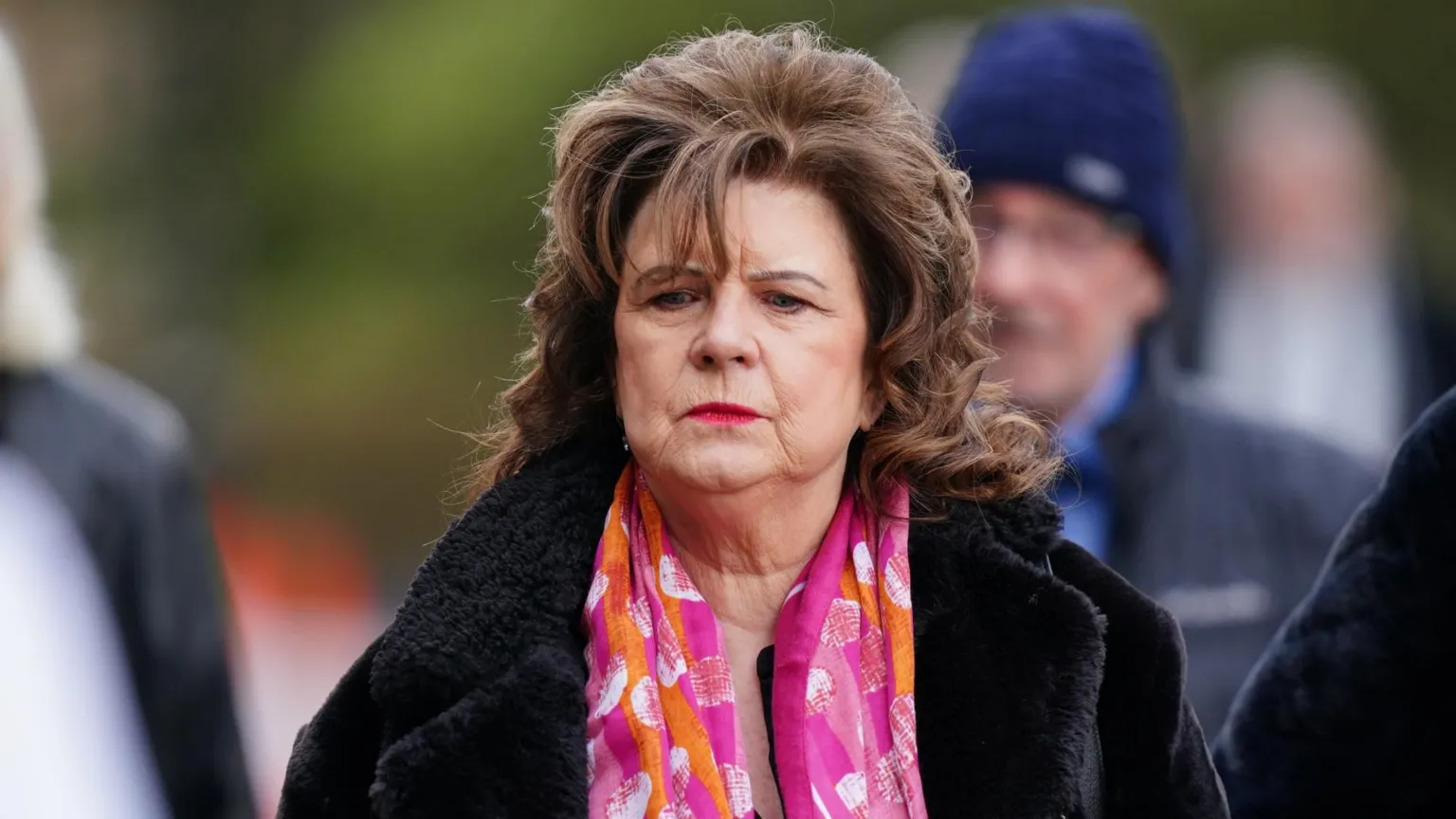  Elaine C Smith who has shoulder, length brown hair is wearning a pink, orange and white silk scarf and a black fluffy coat. She has a solemn look on her face