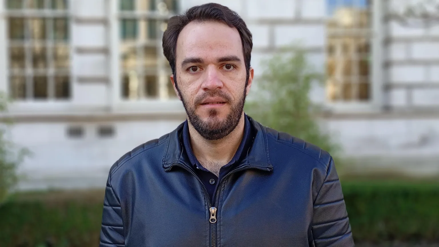 Iman Talebian has short black hair and beard and s looking at the camera, he has a black leather jacket on. He is standing outside the Portland stone building of the Ulster Museum and green shrubbery can be seen behind him.