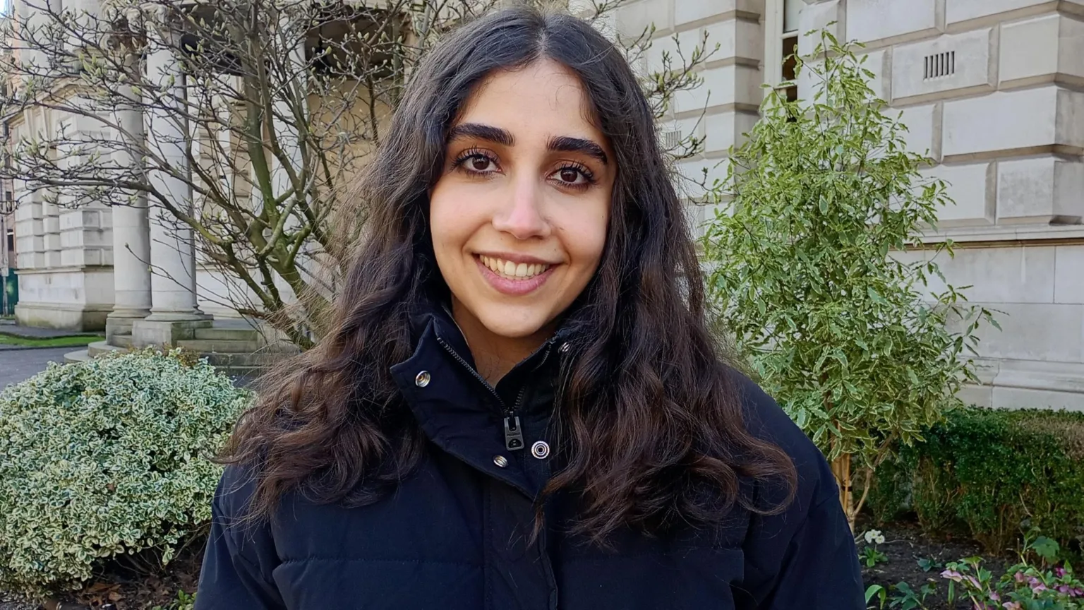 Fatemeh Sadat Mosavi has dark hair and is smiling at the camera, she has a black puffy jacket on. She is standing outside the Portland stone building of the Ulster Museum and green shrubbery can be seen behind her.
