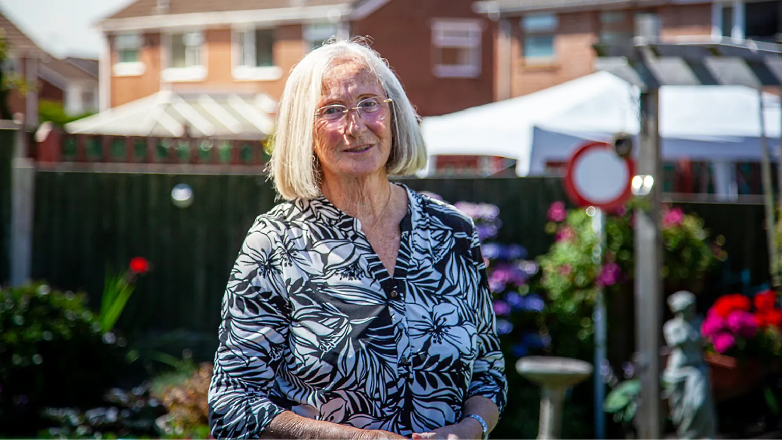 Yeti Television/BBC Cymru Wales Margaret Owen is a white woman in early old age. She has white bobbed hair, wears rimless glasses and a black and white flower-patterned blouse. She is standing outside on a sunny day. The blurred background shows housing, a dark wooden fence and colourful garden plants. She looks directly at the camera and is half-smiling.
