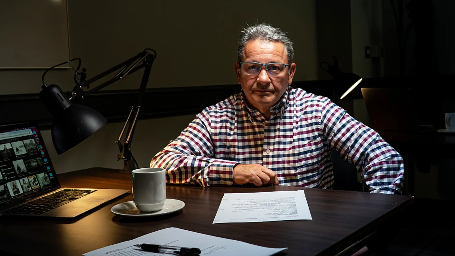 Yeti Television/BBC Cymru Wales Jez Hemming is a white middle-aged main. He has dark greying hair, is tanned and wears grey-framed glasses and a white, black and red checked shirt. He is sitting in a dimly lit room in front of a dark wooden table which has two pieces of paper, a pen, a mug and plate, a laptop and an black anglepoise lamp on it, which is casting light onto the desk. Another lamp is lit in the background.