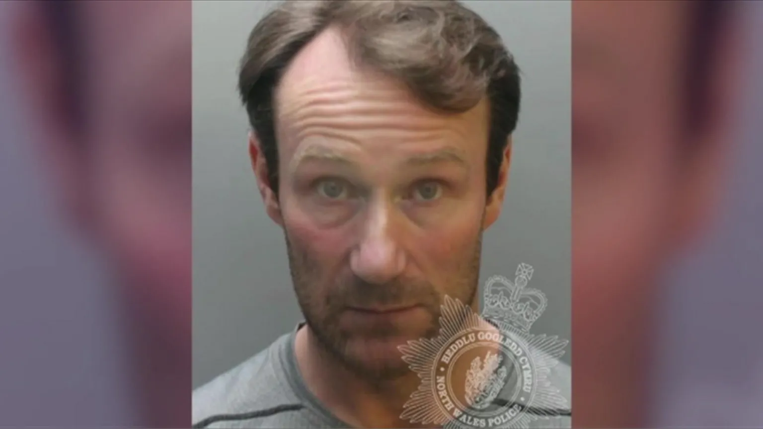North Wales Police Christian Williams is a white man with short light brown hair. He is unshaven and wears a grey top. It is a head and shoulders police custody shot with a plain grey background and a watermark with the North Wales Police/Heddlu Gogledd Cymru crest on the lower right hand side of the photo.