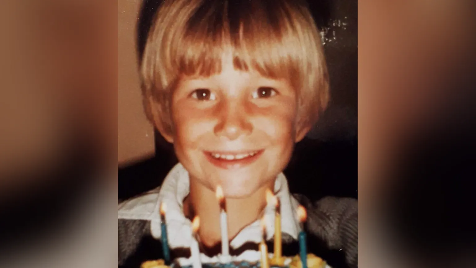 Family photo Andrew Hamilton as a young boy. He has dark blond hair and is wearing a rugby-style top with a white collar. It's a head and shoulders shot of him with a birthday cake in front, with six candles on top. He is smiling directly at the camera.