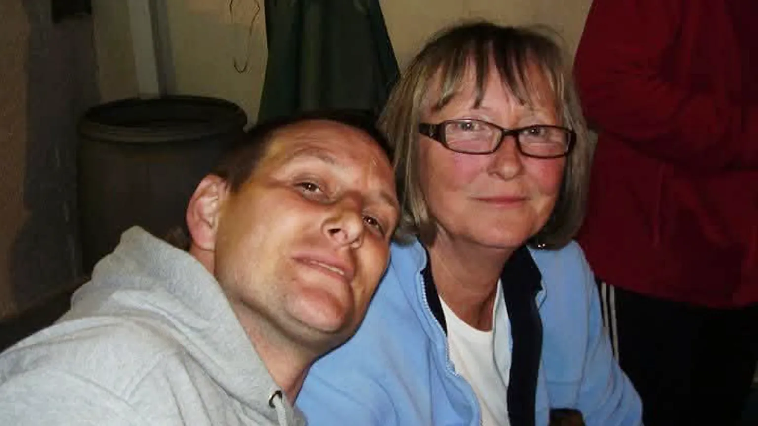 Family photo Andrew Hamilton with his mother Margaret. He is is early middle age with very short cropped dark hair and is tanned. He wears a grey hoodie top. She has dark blonde streaked hair, wears brown rectangular-framed glasses and a white -shirt covered with a light blue fleece jacket. They are sitting down and he is leaning his head on her shoulder. Both look at the camera and are slightly smiling.