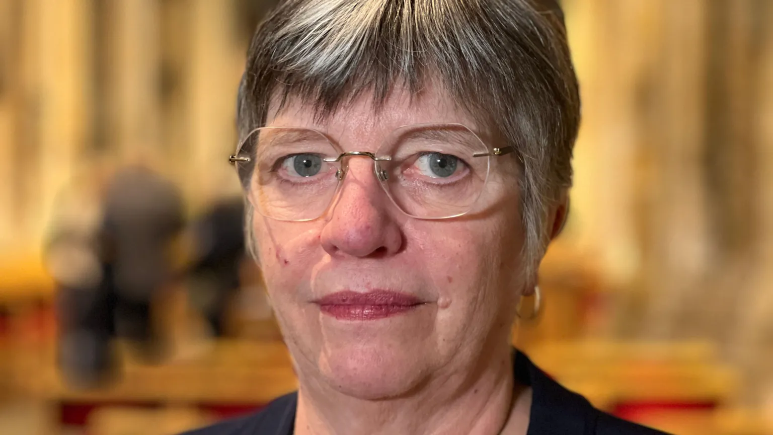 Michelle Lyons/BBC A woman with short grey and black hair wearing frameless glasses. She is standing in a church.