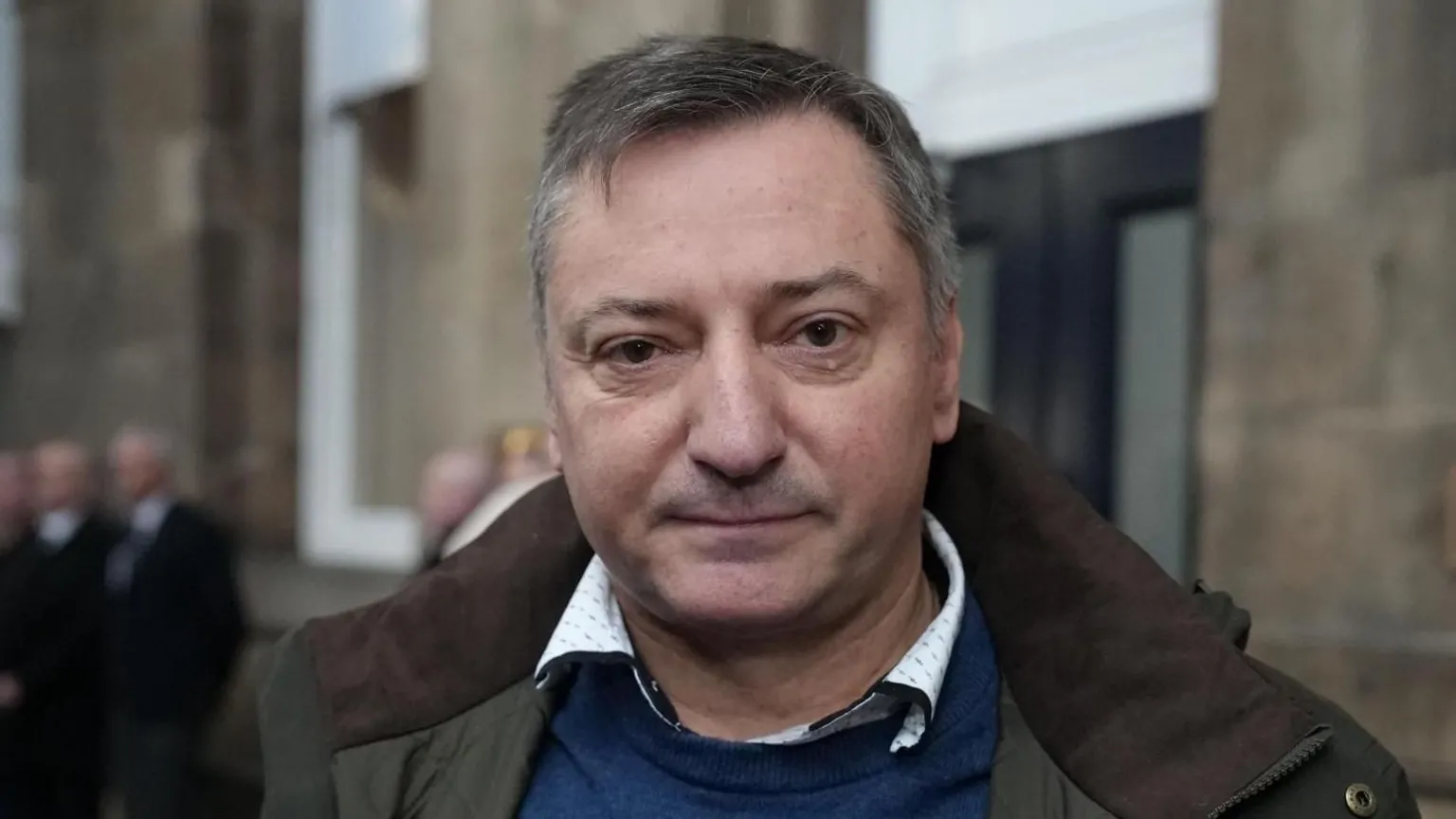 Stuart Prandle/BBC A man with short grey hair wearing a blue jumper and green and brown coat standing on a railway platform