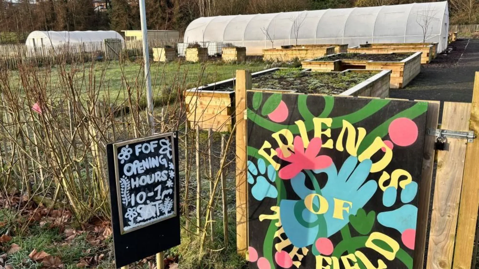 A large community garden. It has a multicoloured hand painted sign that says 'friends of the field' on the front gate. Another black and white sign says 'FOF opening hours 10-1pm Sat'. There are two large white polytunnels and wooden raised beds in the garden.