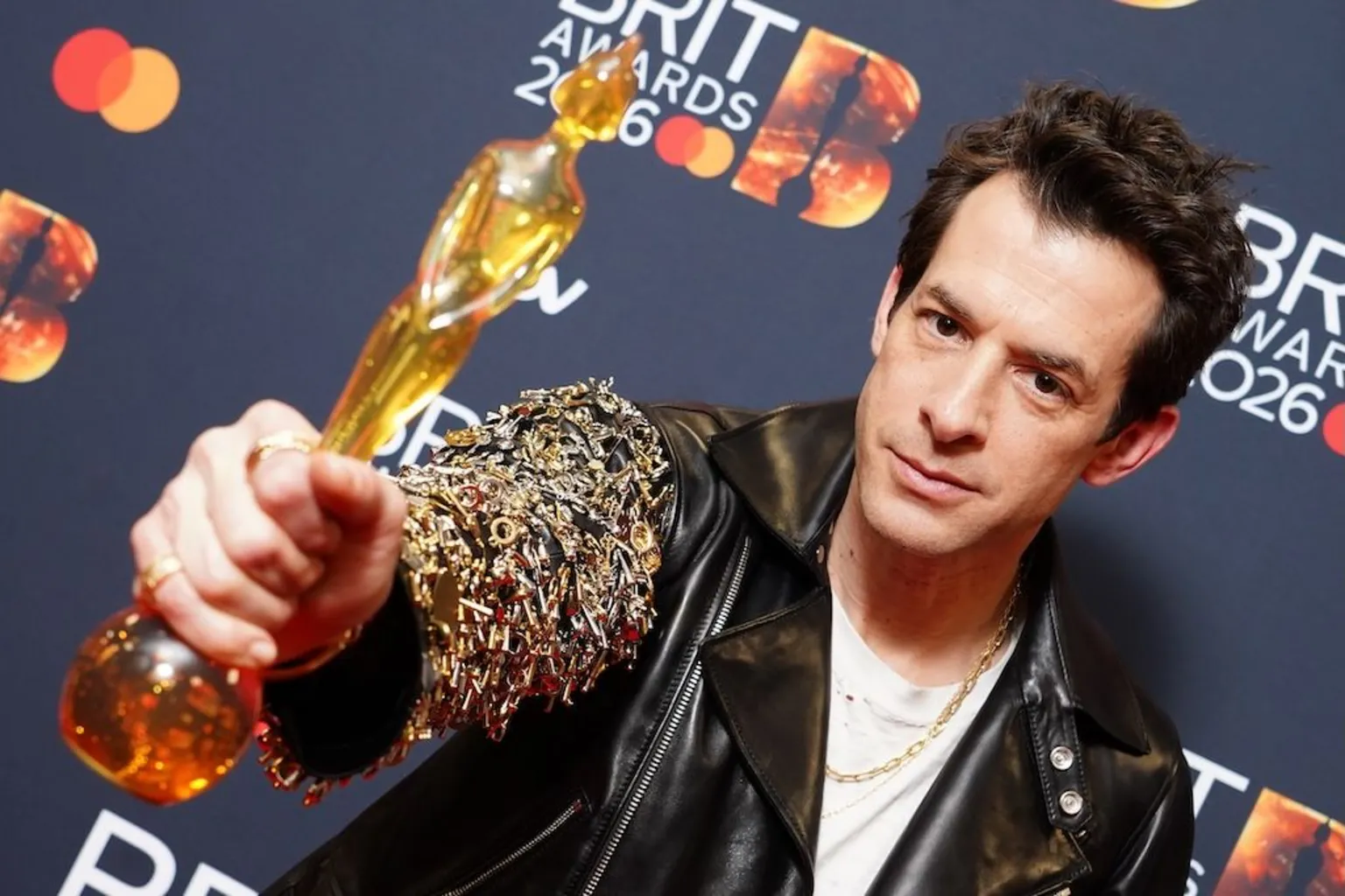  Mark Ronson holds his award up to the camera. He is wearing a leather jacket from the Celine collection.