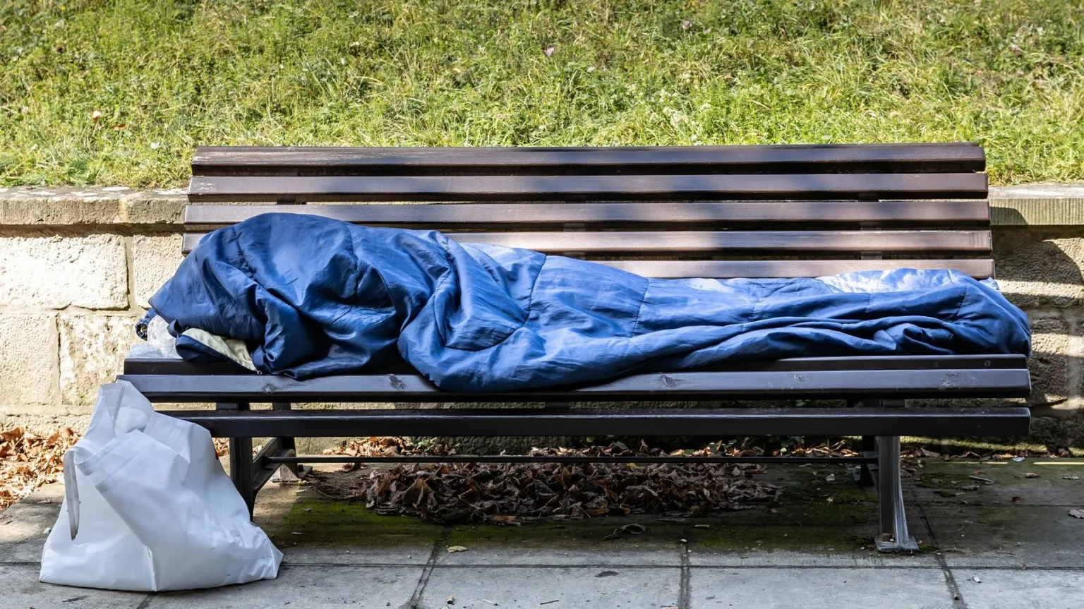 Getty Image A blue sleeping bag style covering on a brown park bench, indicating homelessness. There is a white bag to the left.