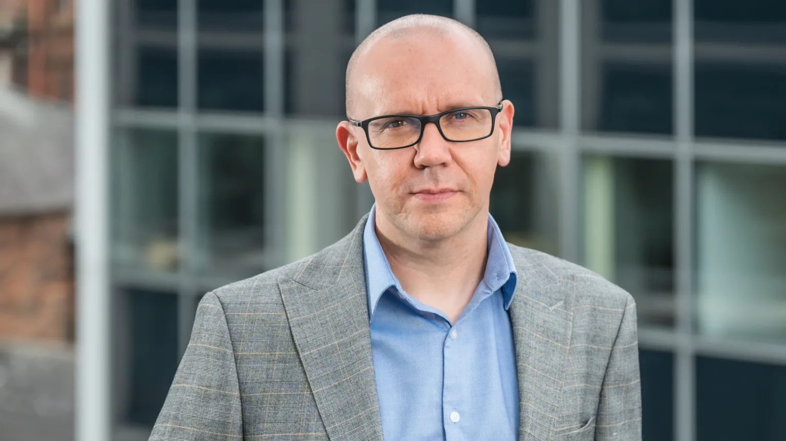 Simon Community A bald man with black square rimmed glasses looks into the camera. He is wearing a light grey suit and a light blue shirt with an open collar.
