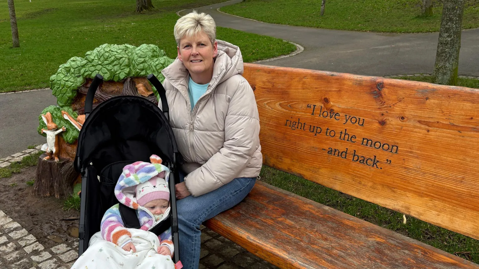 Roberta Carser, a woman with short, blonde hair, sits on a carved wooden bench with her hand on her granddaughter's buggy. She is wearing a padded beige jacket, blue jeans and a light blue t-shirt. Her granddaughter Torie is wearing a multi-coloured hooded jacket and a pink and white woolen hat. The bench features decorative carvings of trees and hares and it is engraved with a line from the book: 