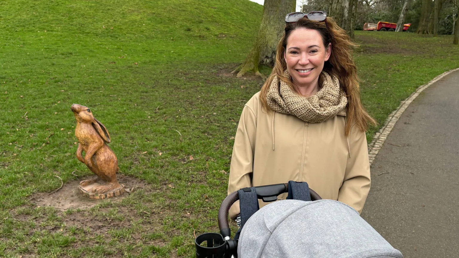 Katie Alexander, a woman with long, light brown hair, smiles at the camera in Wallace Park in Lisburn. She is pushing her daughter's grey and black buggy which is partially visible in the photo. Katie is wearing a beige coat, a matching woollen scarf and she has large sunglasses on top of her head. There is a small wooden statue of a hare to her left and a large tree behind her. 