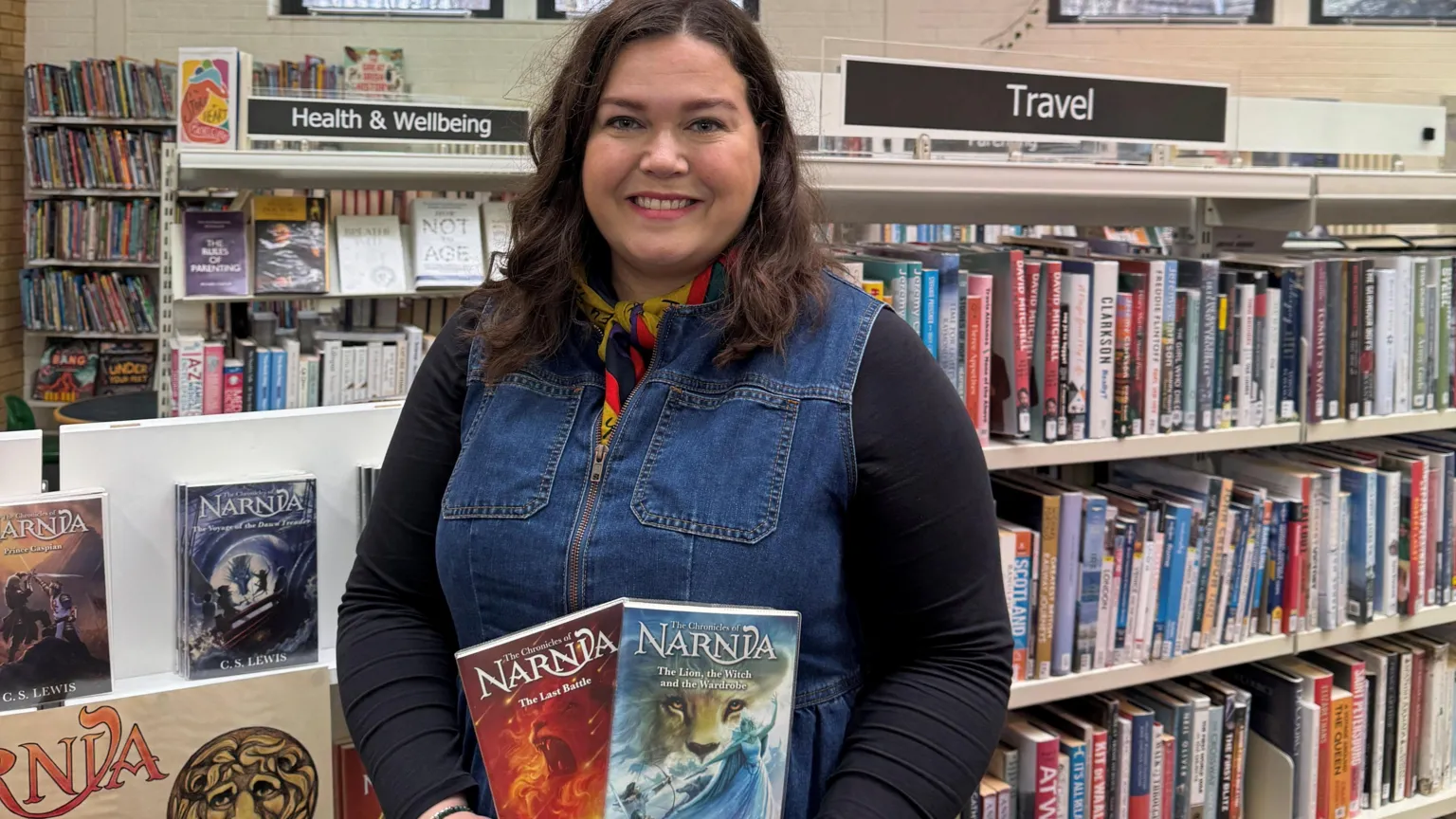 Woman in denim, standing holding two books. She is in a library. 