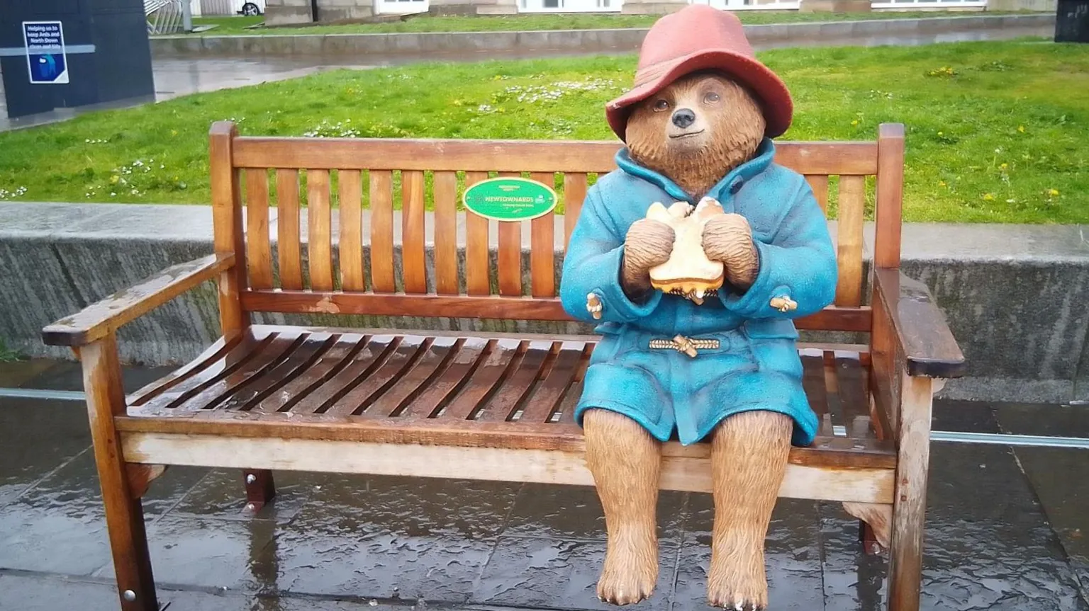 A statue of Paddington Bear sitting on wooden bench in a park. He is wearing a blue duffle coat and a red hat. He is eating a marmalade sandwich.