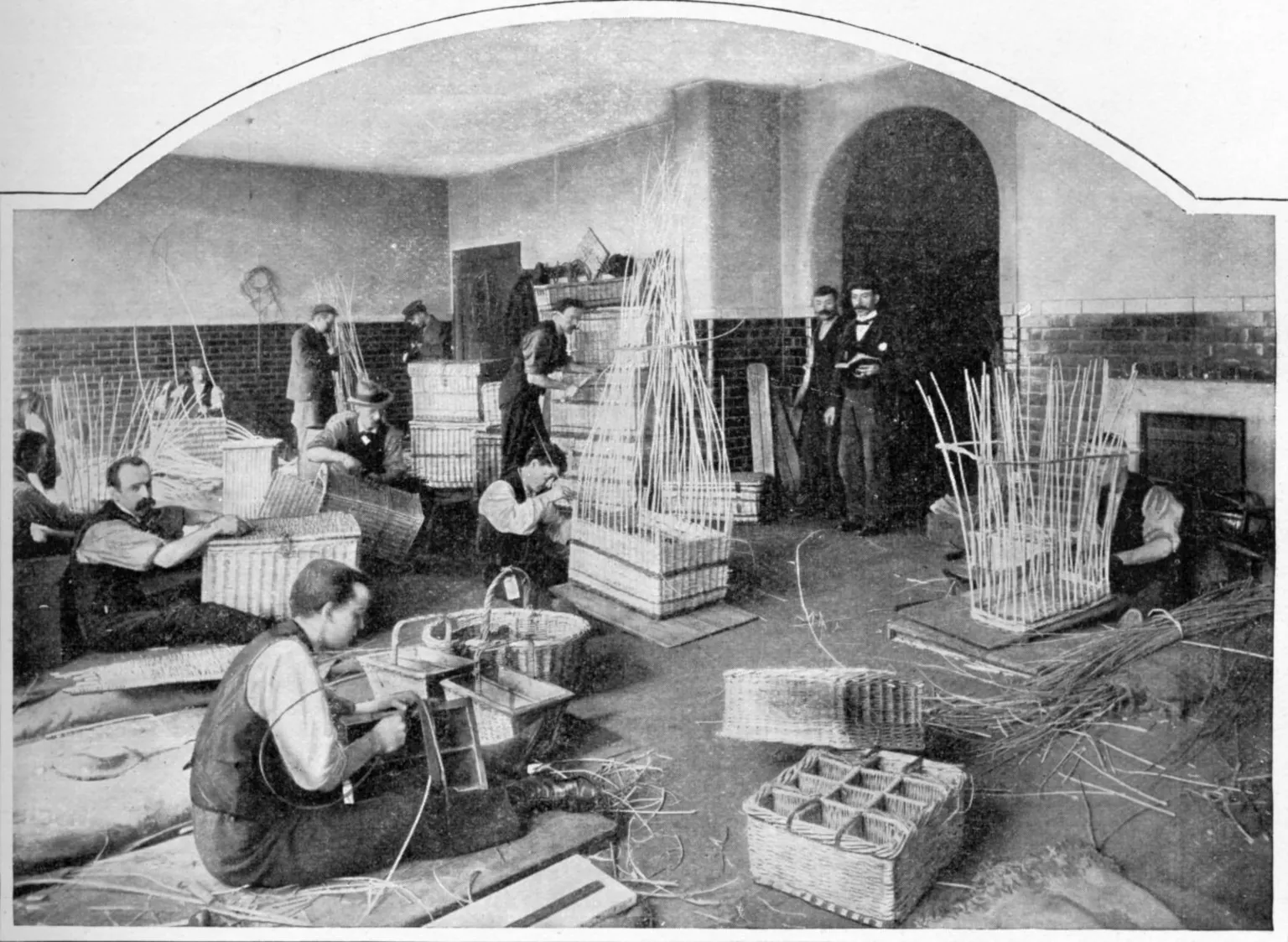 A black and white photograph showing blind men in waistcoats sat on the floor a factory warehouse weaving baskets. Others are stood in the distance working on the baskets. Two men in three piece suits stand in an archway looking at the work.