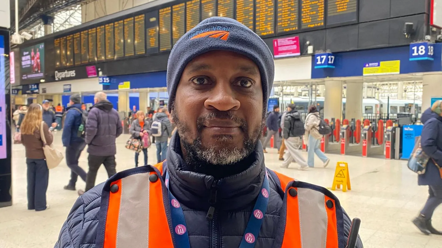 A medium close up of Yusuf Hussein stood at a busy station looking into the camera. He is wearing an orange high-vis jacket, a lanyard for his job, a navy blue jacket, and a blue Network Rail beanie hat. Behind him, you can the departures and arrivals screens, barriers for platforms, and lots of people walking in different directions. 