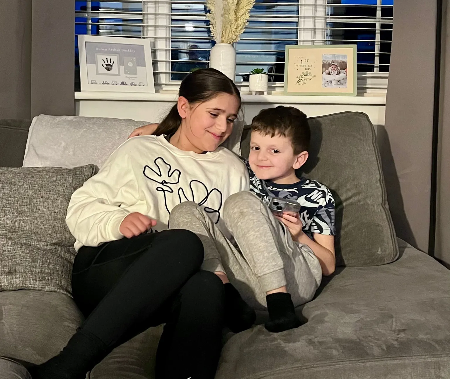 Olivia and Arlo sit close together on a grey sofa, smiling. Arlo has his arm around his sister. 