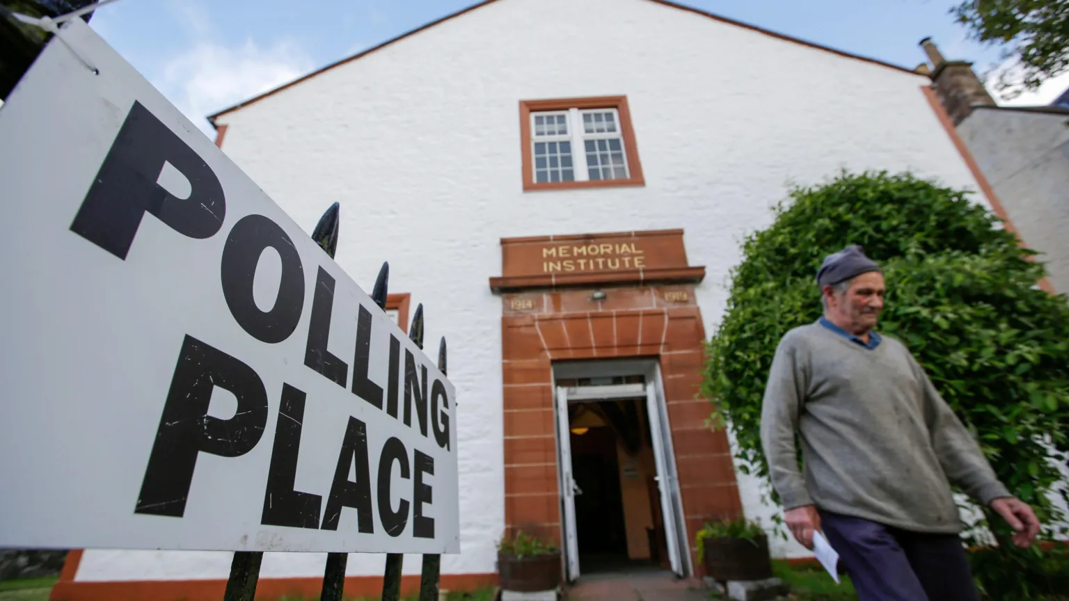  A man wearing a grey jumper and a blue hat leaves after casting his vote at the Moniaive polling station in Dumfries and Galloway. The building has white walls and the doorway is made of red sandstone. A sign on the left of the image says Polling Place.