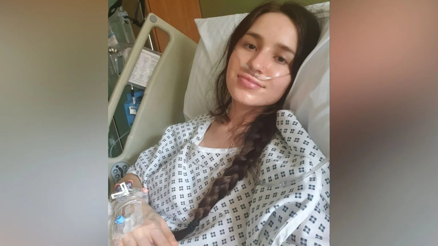 Amy Peckham-Driver Amy Peckham-Driver in a selfie-style image as she smiles at the camera while lying in a hospital bed. She has long brown hair which has been tied into a plait that rests over one of her shoulders and her torso. She wears a white hospital gown with blue squares on it. She has various medical tubes connected to her hand as well as a tube around her face and nose.