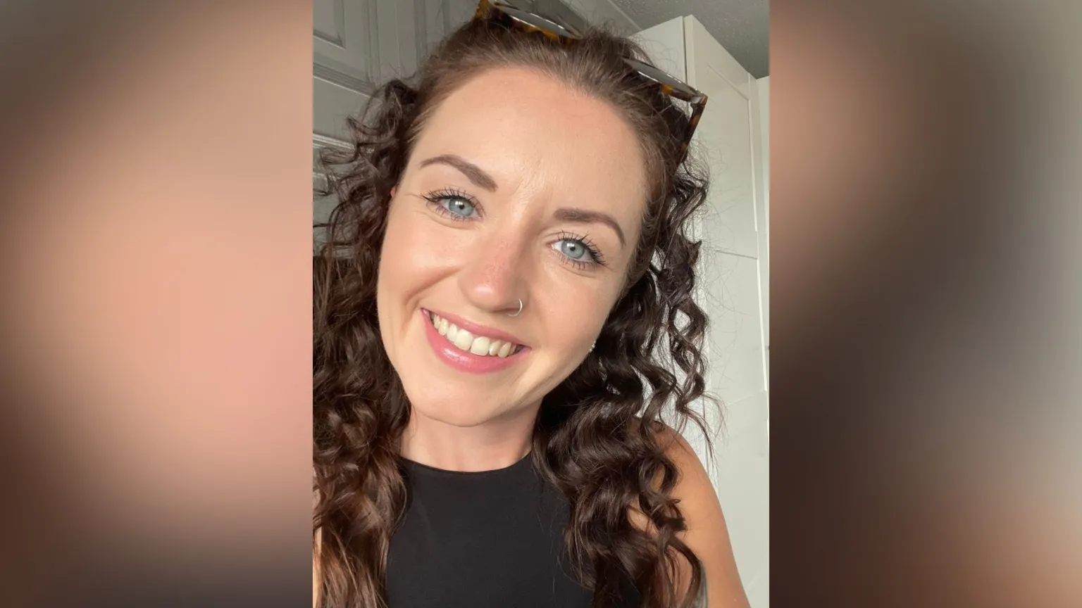 Faye Ramsey A selfie-style image of Faye Ramsey who is smiling at the camera. She has long brown hair which is curly and also has a nose ring on her right hand nostril. Her glasses rest on the top of her head and she is wearing a black sleeveless top.