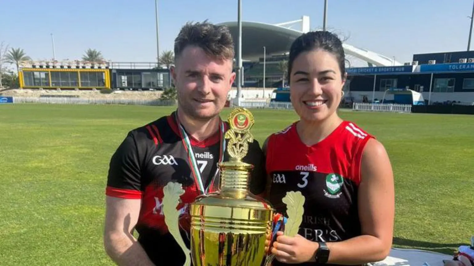 Conor Duggan Conor Dugganand woman in GAA sports tops are holding a gold trophy together. They are standing outside on a field and buildings can be seen in the background. Conor has short brown hair and is wearing a black GAA top with a red stripe across the centre. The woman has black hair pulled back behind her head and is wearing a red and black GAA sleeveless jersey. 