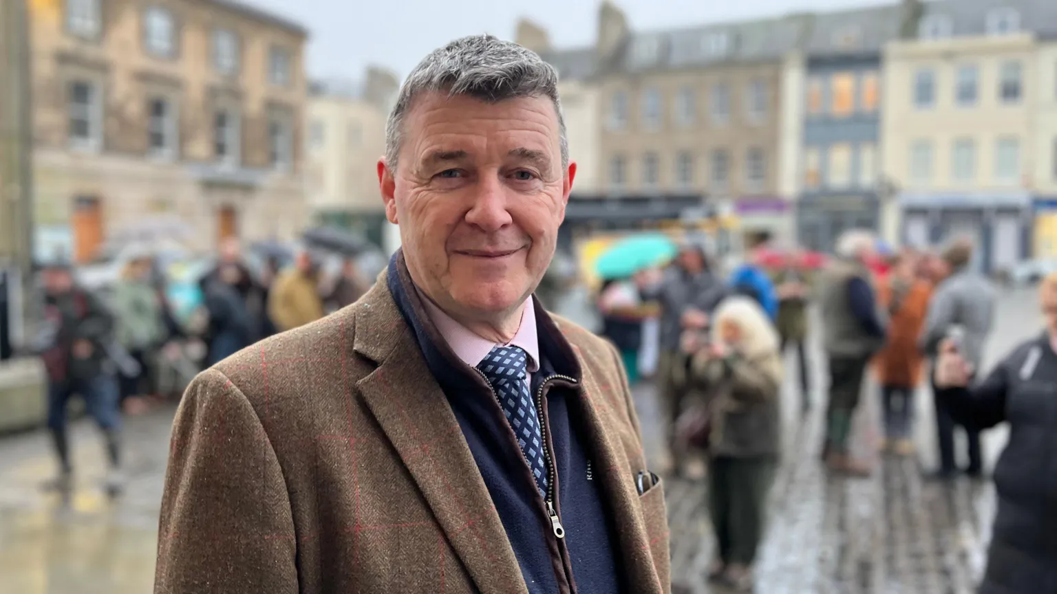 A grey-haired man with a brown jacket, pink shirt and blue tie faces the camera with a busy street scene behind him
