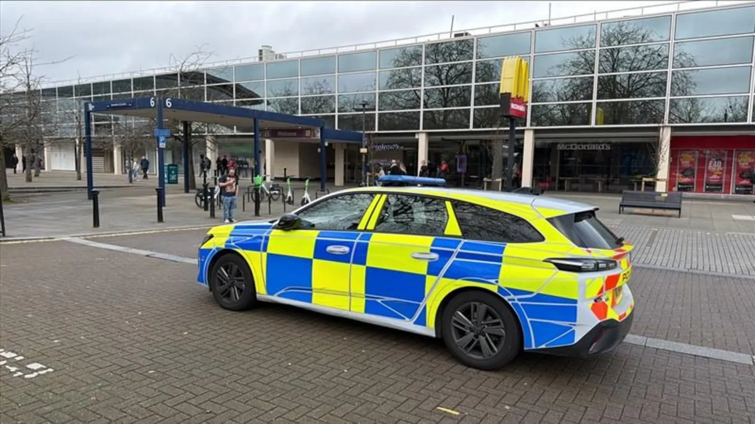 Richard Knights/BBC A marked yellow and blue police car is parked outside Centre:MK - which is a glass building with a yellow McDonald's sign outside. It is daytime and it is cloudy above.