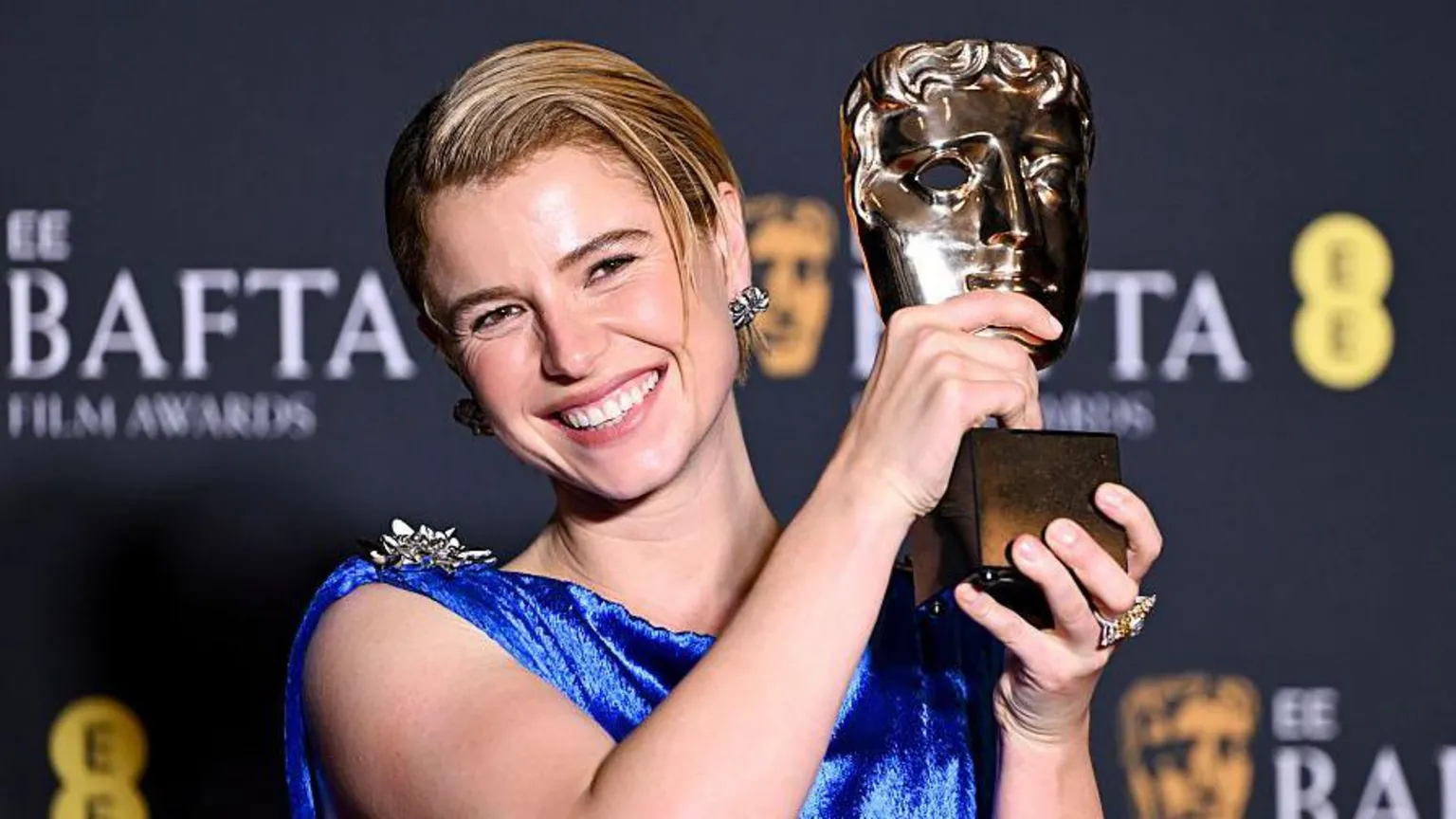  Buckley has blonde hair which is tied back, she is holding up her BAFTA award and is wearing a blue dress.