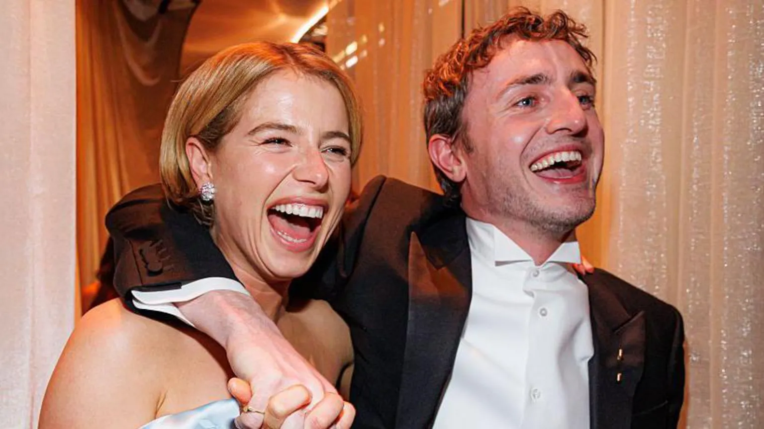  Buckley with short blonde hair, diamond earring and off the should dress is smiling and she walks along with Paul Mescal. His arm is around her shoulder as he smiles. He is wearing a black suit jacket, white shirt and has short dark hair. 