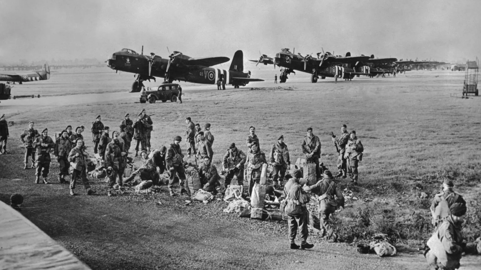  An old black and white photo from September 1944 during World War Two. There is a group of soldiers on a field preparing their parachutes and equipment before taking flight for Operation Market Garden in the Netherlands. There are several planes in the distance.