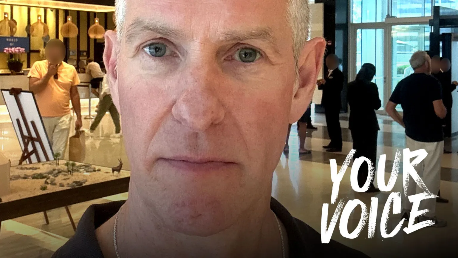 Ian Scott A man looks serious while standing in front of people milling about an airport shopping area.