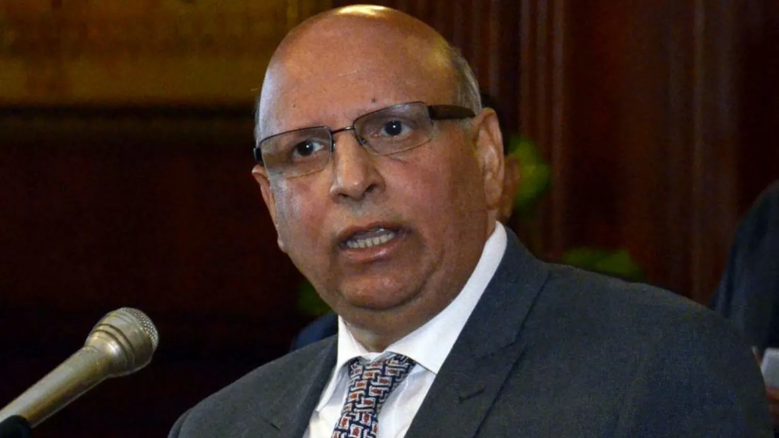  Mohammad Sarwar, who is bald with glasses, speaks into a microphone. He is wearing a dark suit, white shirt and patterned tie. 