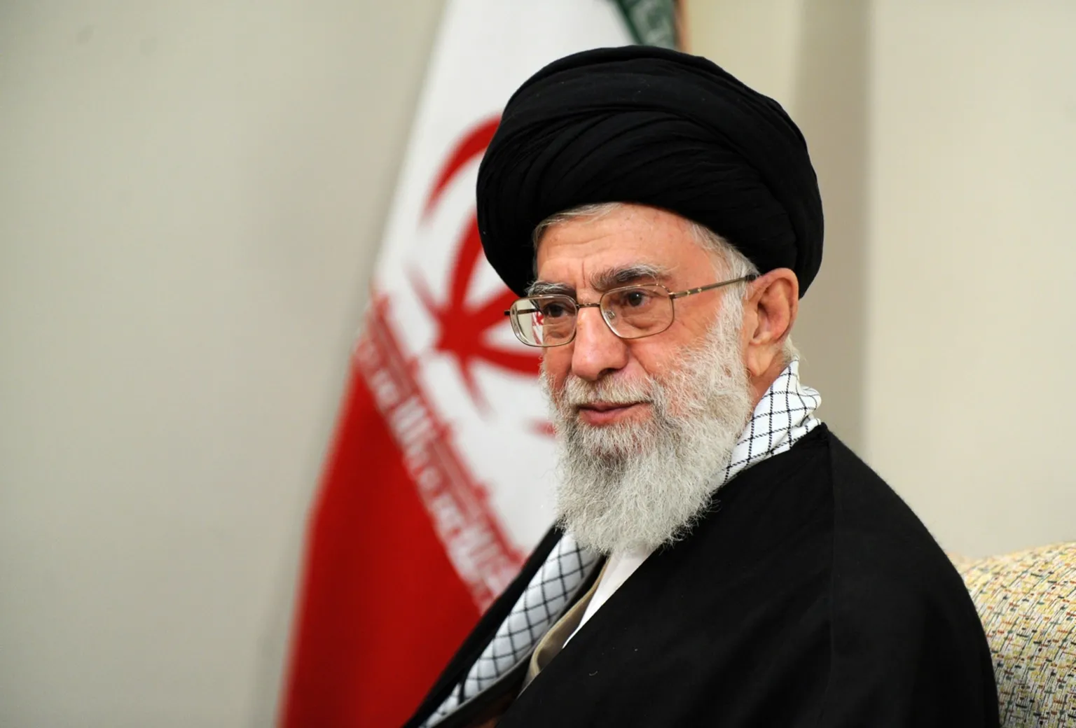  Ayatollah Ali Khamenei, who has a long grey bears an glasses, sits side on in front of a wall and a large flag. He is wearing a black robe and head piece. 
